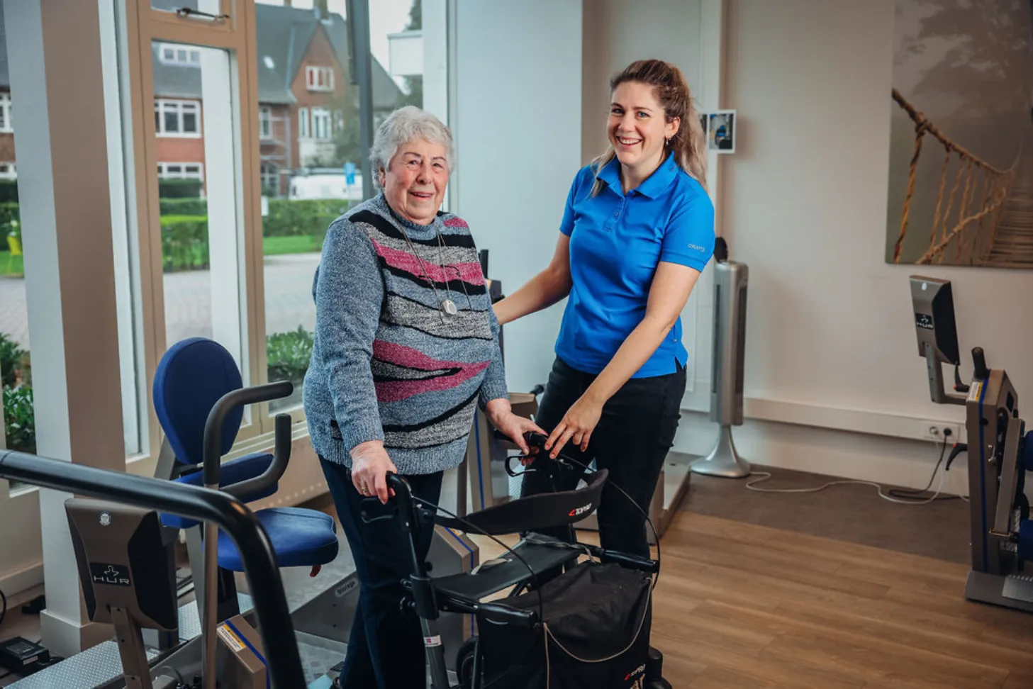 Een oudere mevrouw staat achter haar rollator, bij een trainingsapparaat. Ze wordt begeleid door een medewerkster van Zorgbalans.